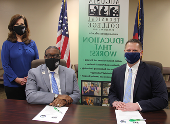Caucasian male wearing a blue face mask, blue suit jacket, white collared shirt, light blue tie, hands folded while sitting down at a table, white paper in front of him with a green pen; African American male wearing a black Augusta Tech branded face mask, light gray suit, silver Augusta Tech lapel pin, white collared shirt, blue/purple tie, silver watch, hands folded white sitting at a brown table, white papers on brown table in front of him with a green pen; Caucasian female standing wearing a black Augusta Tech branded face mask, blue shirt, hands folded together, black pants, background features an American flag, the state of Georgia flag, large retractable banner showing old Augusta Tech logo, verbiage reads: eg8.joyerianicaragua.com, Education That Works; bulleted list reads Allied Health Sciences and Nursing, Business and Personal Services, and Engineering Technology, Industrial Technology, and Learning Support, photos underneath the bulleted list.